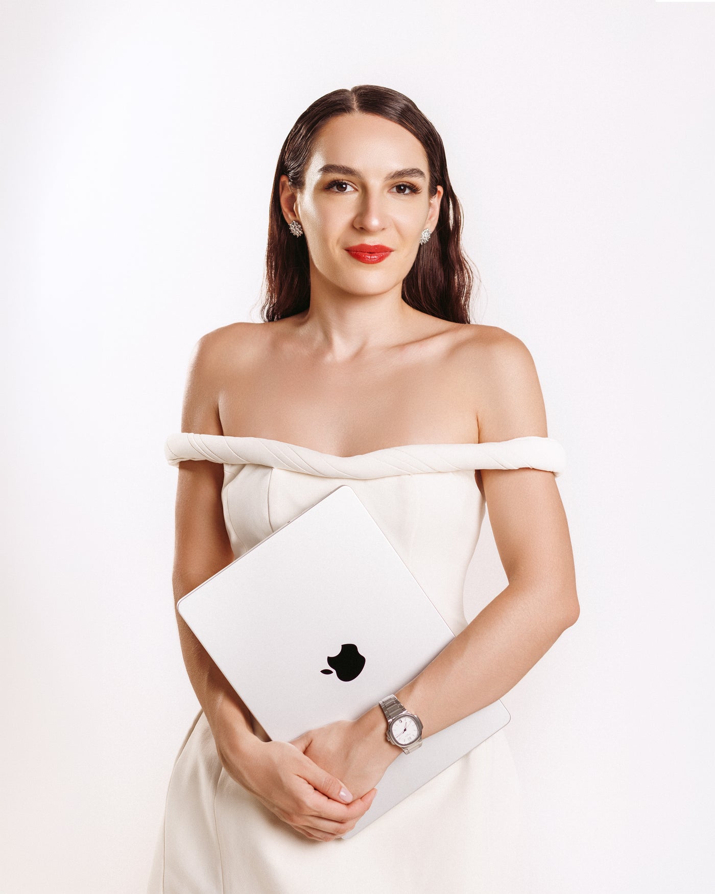 A woman with dark hair wearing a white off-the-shoulder dress holds a silver Apple laptop against a plain white background.